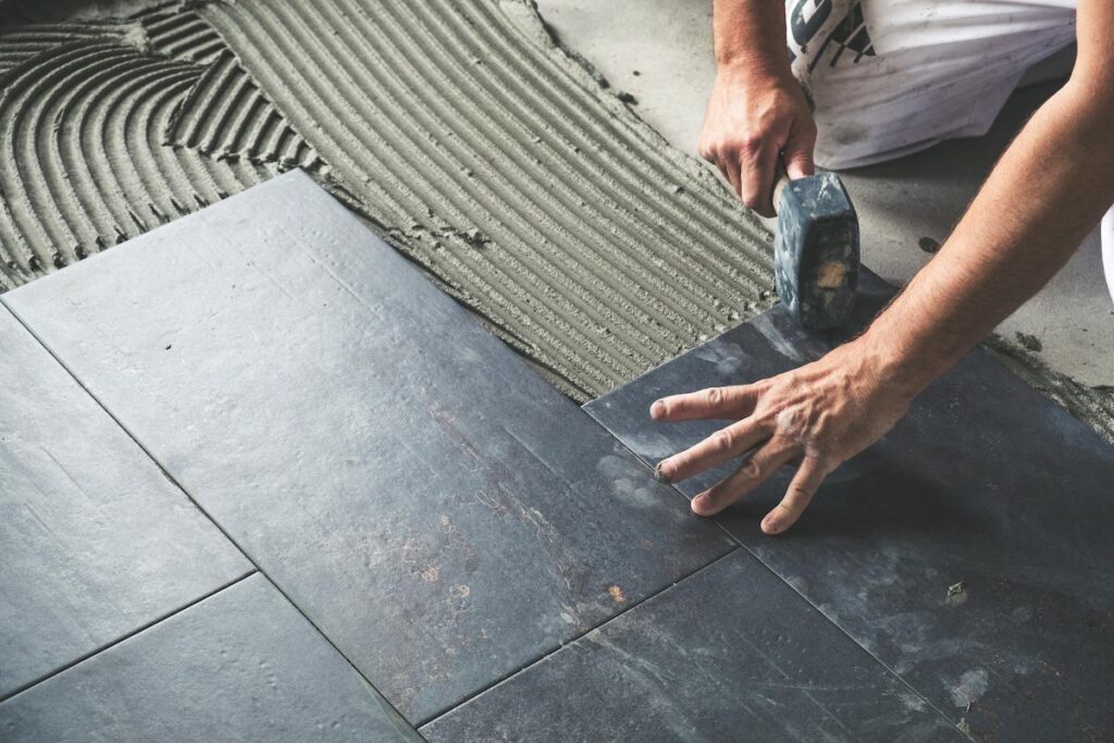Worker placing ceramic floor tiles on adhesive surface, leveling with rubber hammer